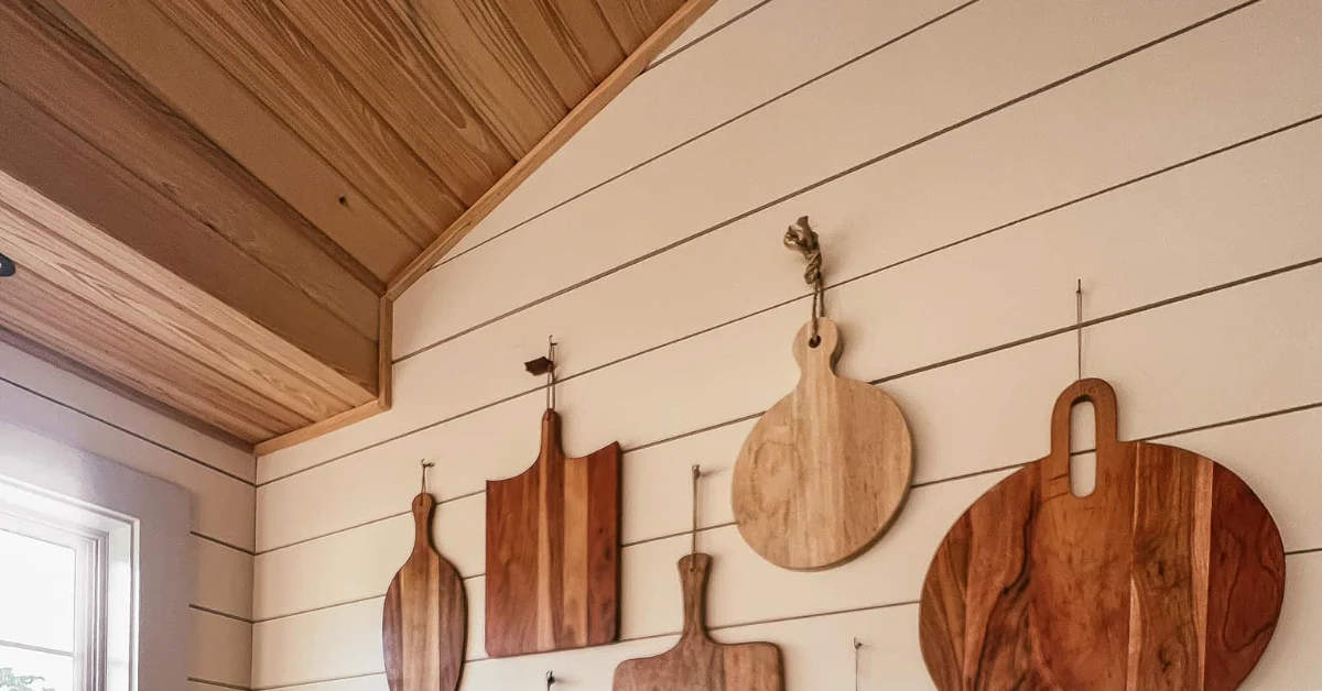 Wooden cutting boards of varying shapes and sizes hanging on a white shiplap wall beneath a vaulted wood-paneled ceiling.