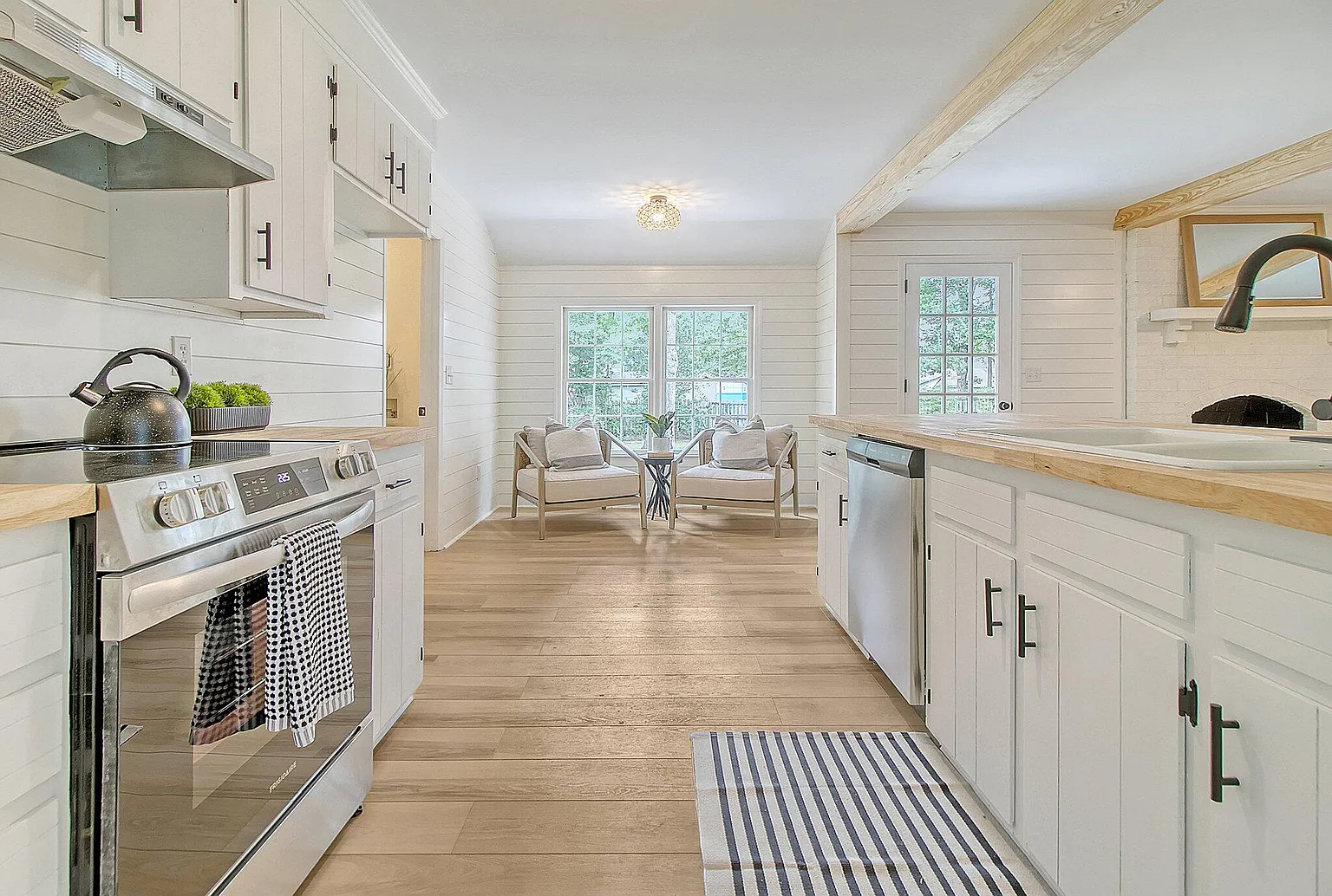 White farmhouse kitchen with shiplap walls and butcher block counters, opening to a sitting nook by large windows.