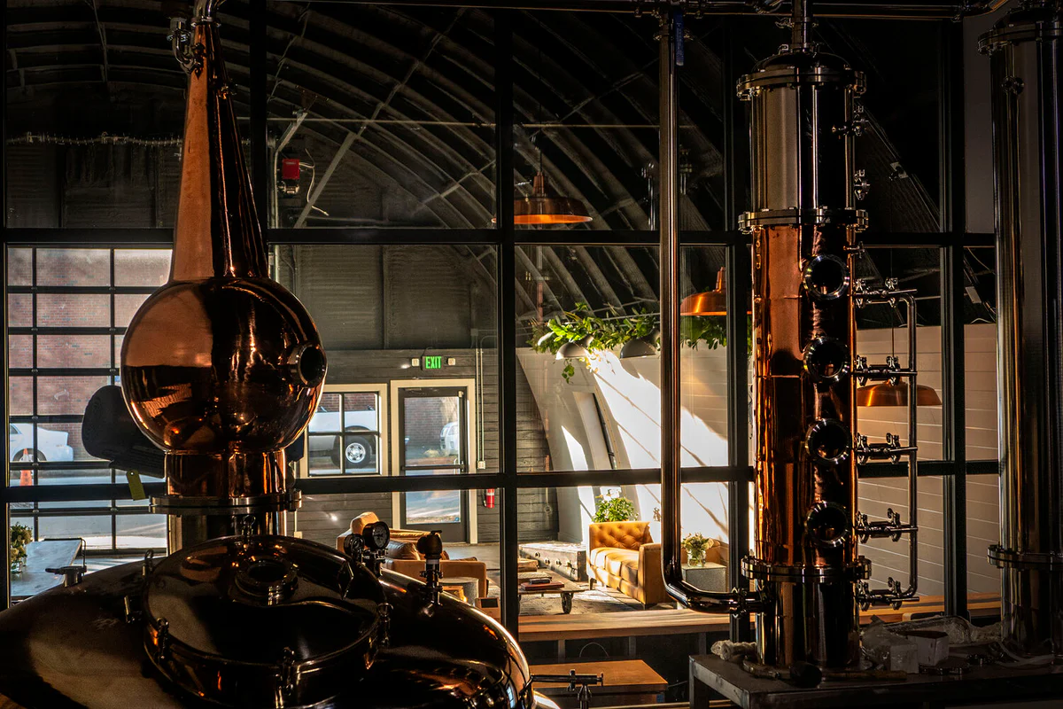 Copper distillery still equipment in the foreground, with a view through glass panels into a lounge seating area beyond.