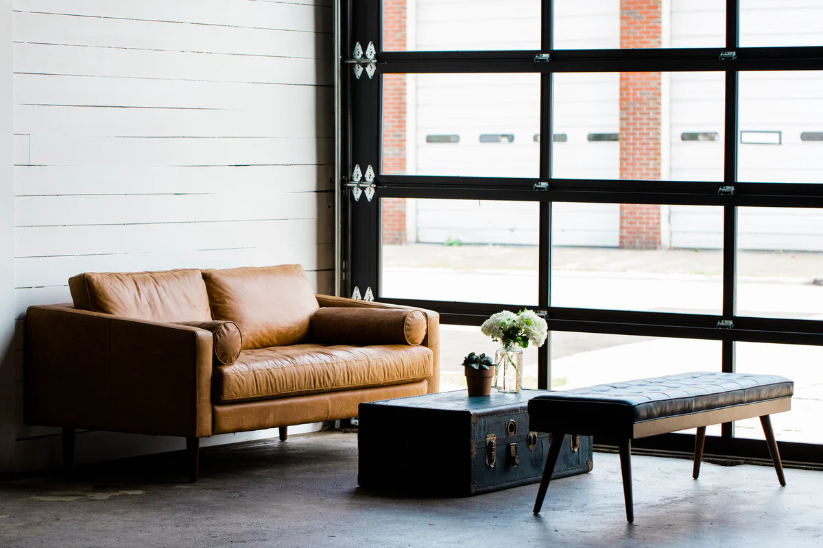 Industrial-style lounge area with a tan leather loveseat, vintage trunk coffee table, and tufted bench beside a glass garage door.