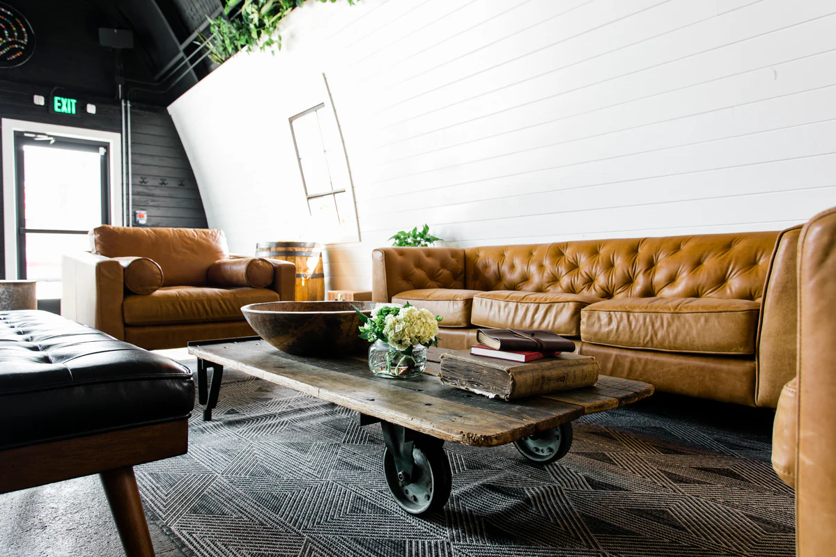 Lounge area of a distillery with tufted leather sofas and a chair, an industrial cart-style coffee table, and a curved white shiplap wall under exposed dark rafters.