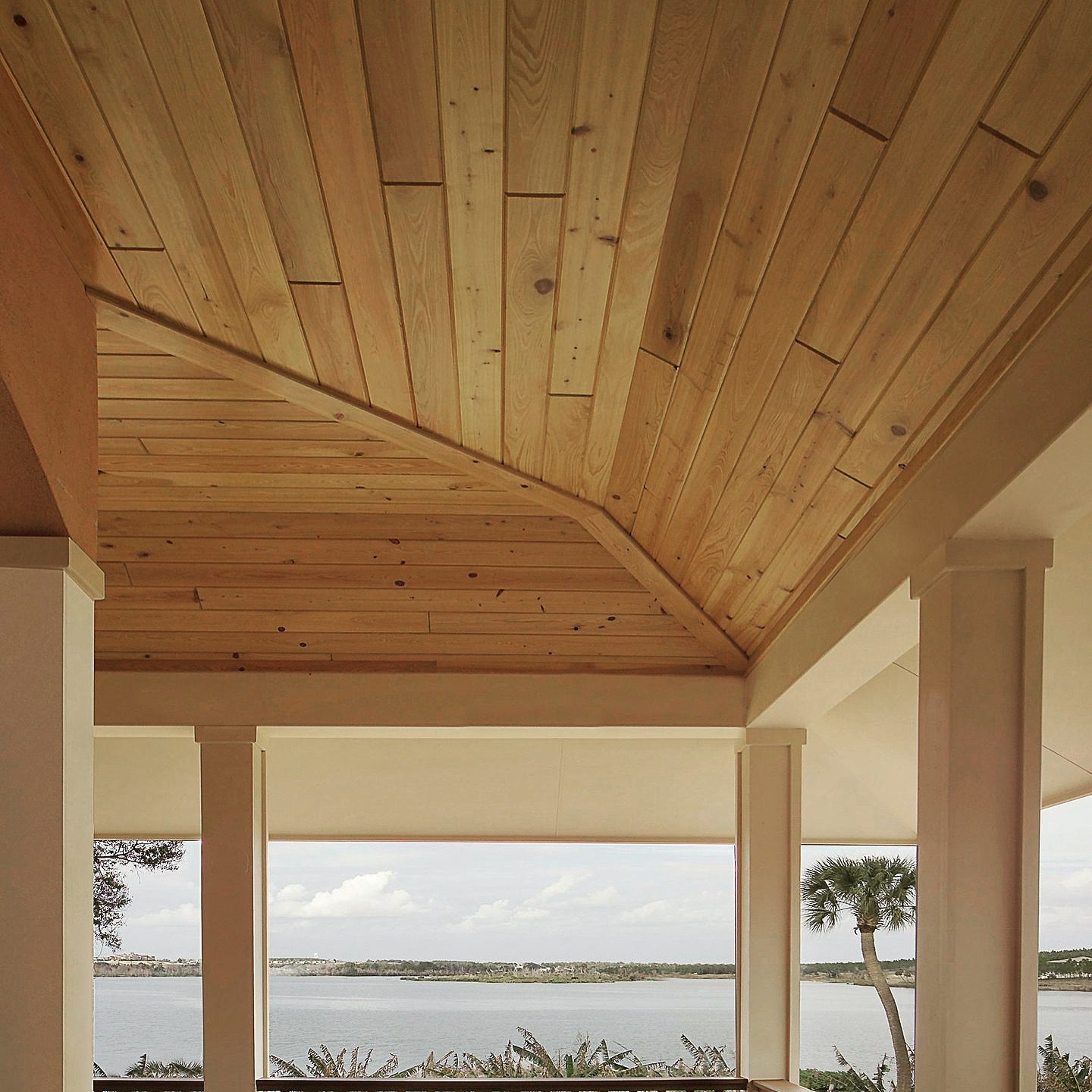 Covered porch with a vaulted wood-plank ceiling and columns, overlooking a waterfront view with palm trees.
