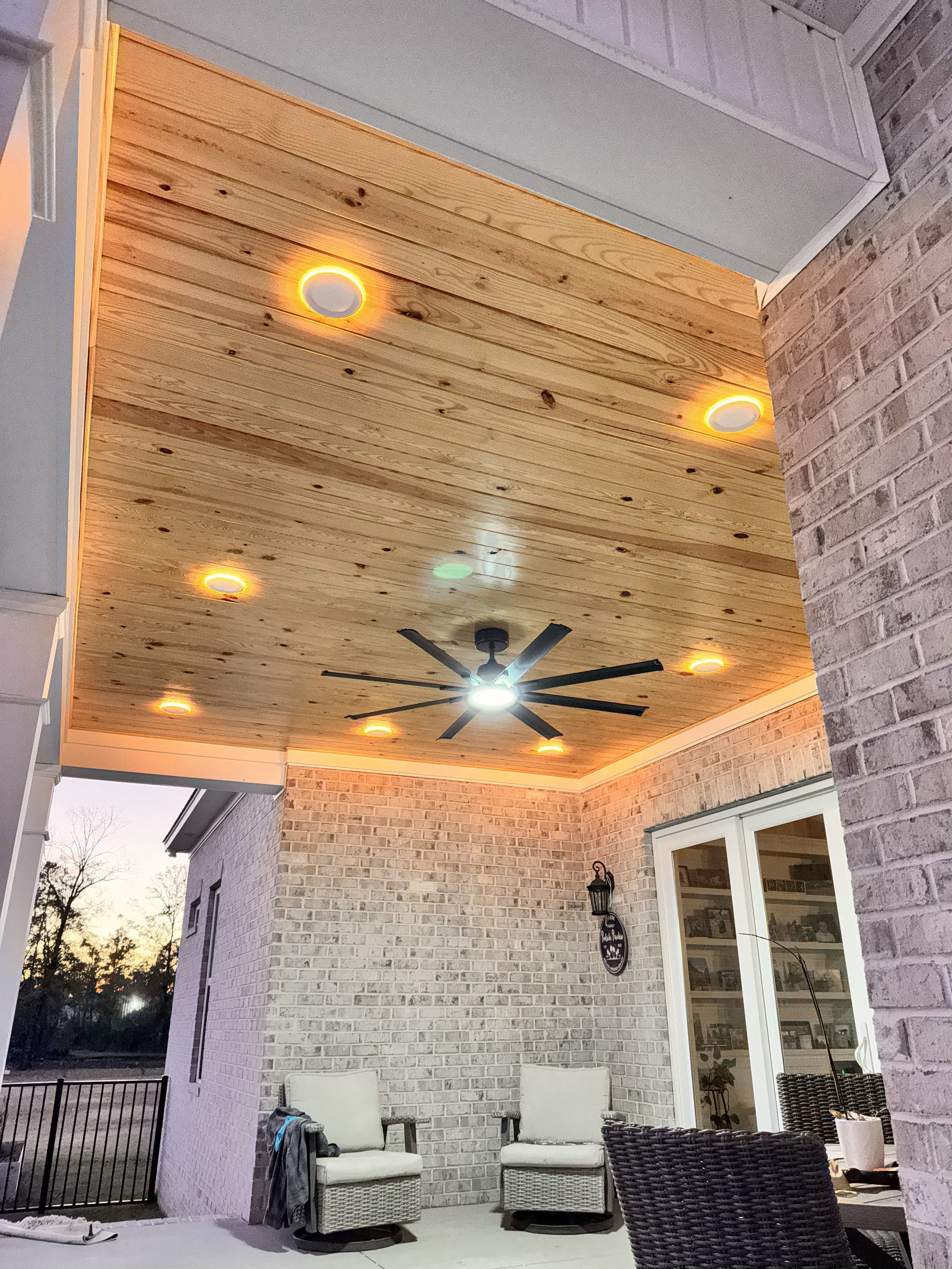 Covered patio with a wood-plank ceiling, recessed lighting, and a ceiling fan, above wicker seating against a brick exterior.