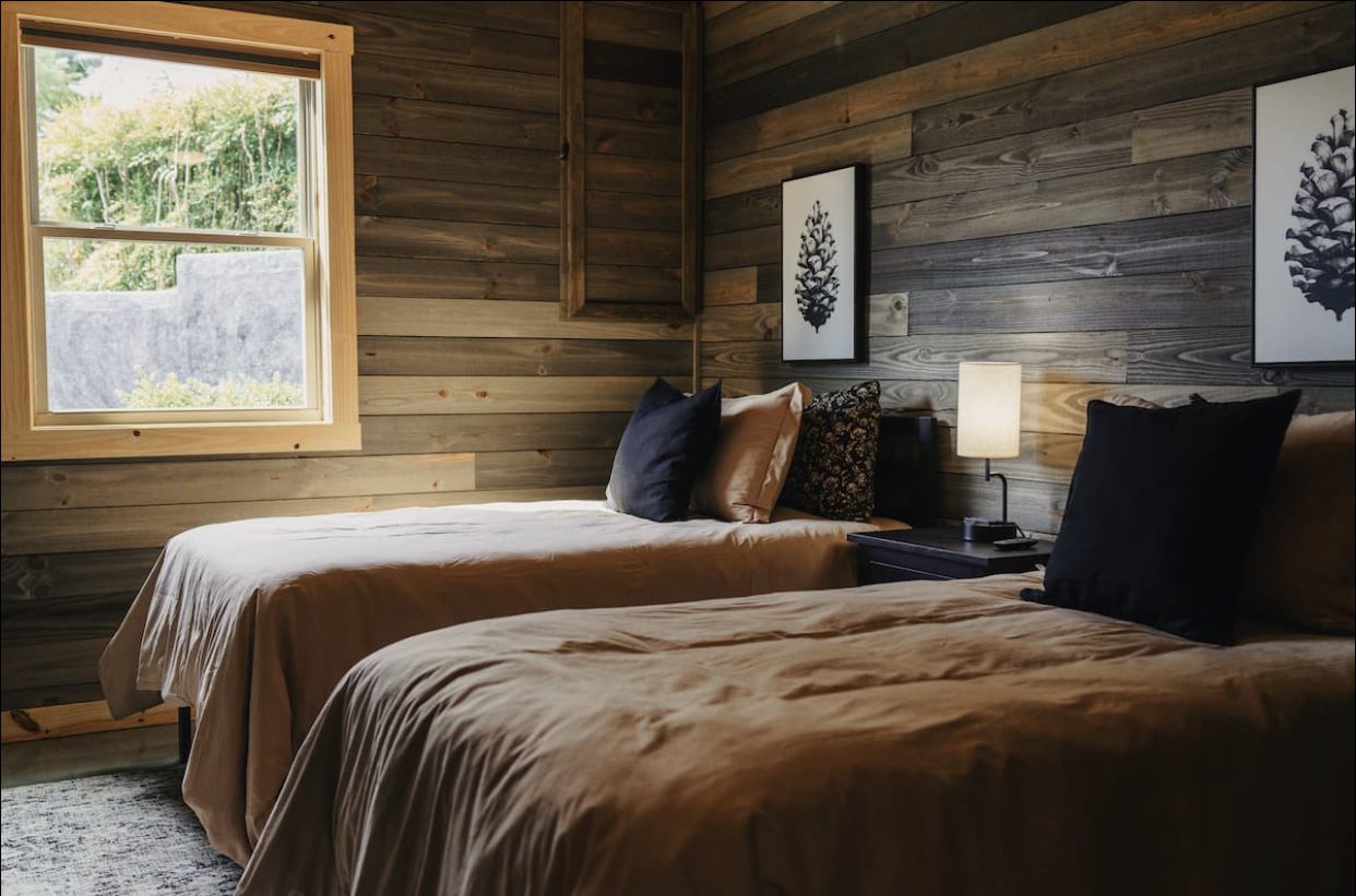 Cabin bedroom with two beds in tan bedding, dark wood-plank walls, pinecone artwork, and a window with a wooded view.