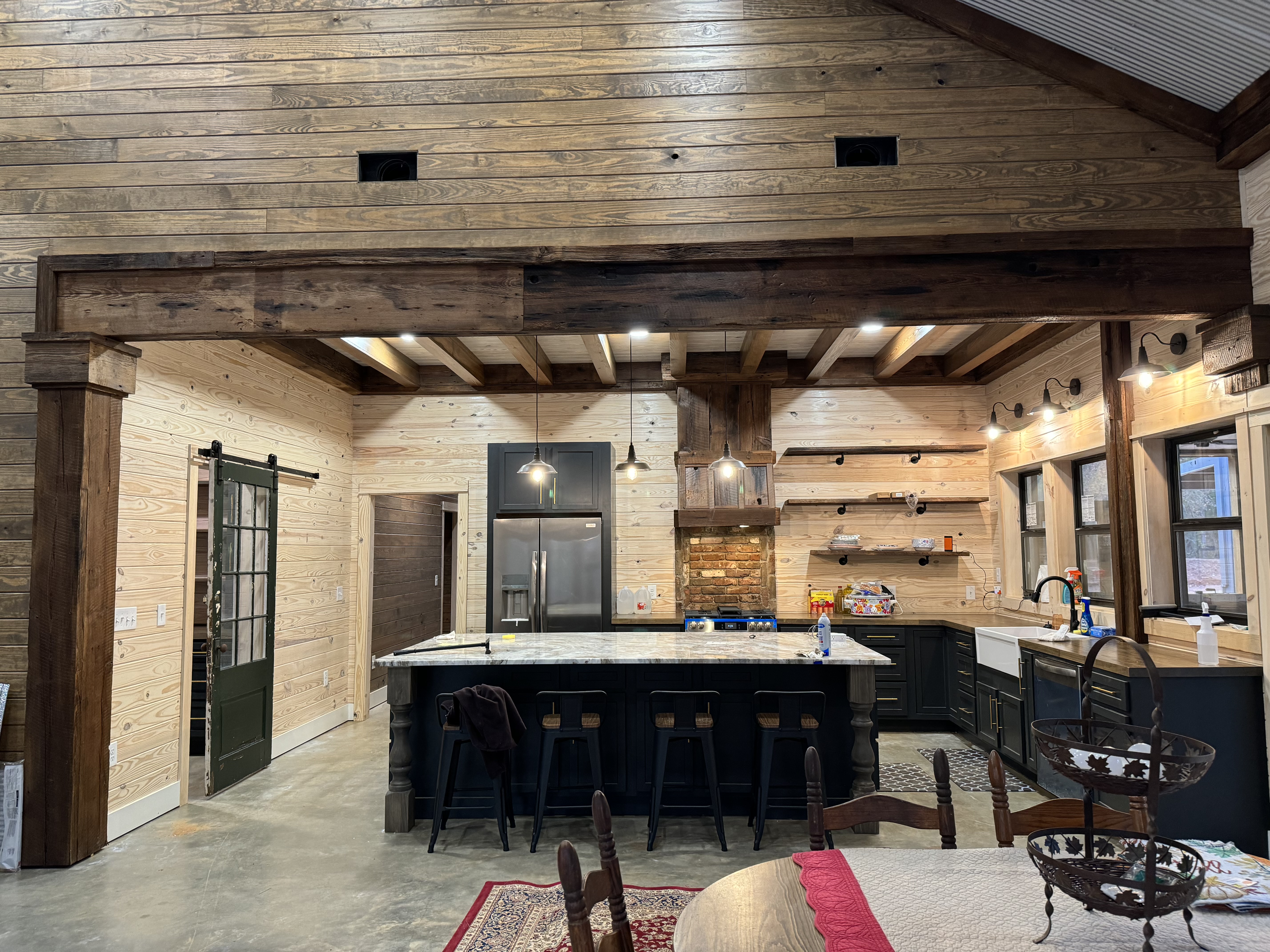 Rustic farmhouse kitchen with a black marble-topped island, exposed wood beams, shiplap walls, exposed brick backsplash, and concrete floors.