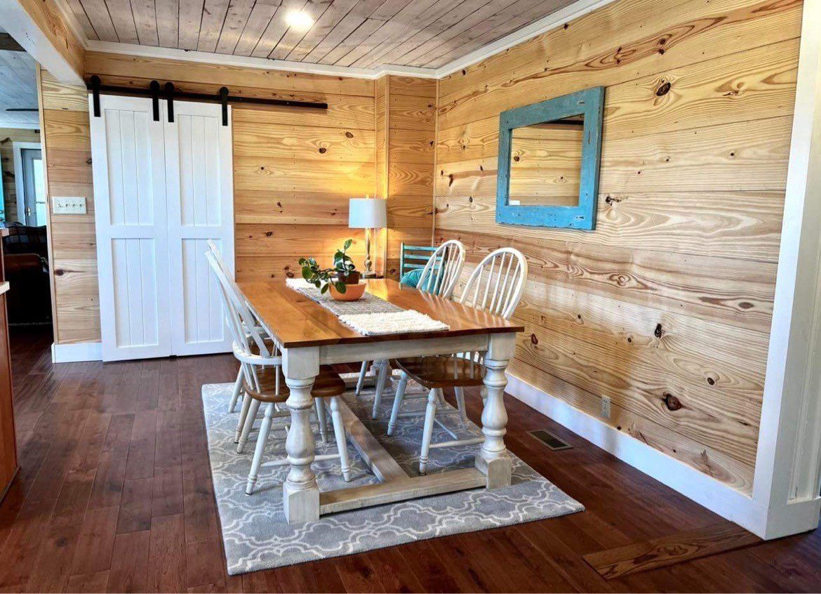 Farmhouse-style dining room with a wood table, mismatched white chairs, and knotty pine walls and ceiling, beside a white barn door.