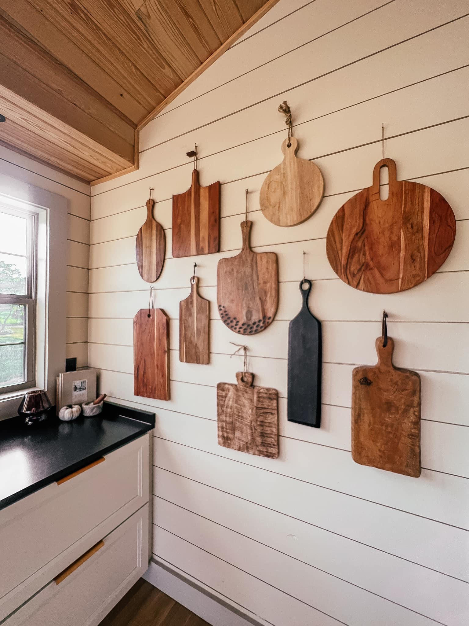 Wooden cutting boards of varying shapes and sizes hanging on a white shiplap wall beneath a vaulted wood-paneled ceiling.