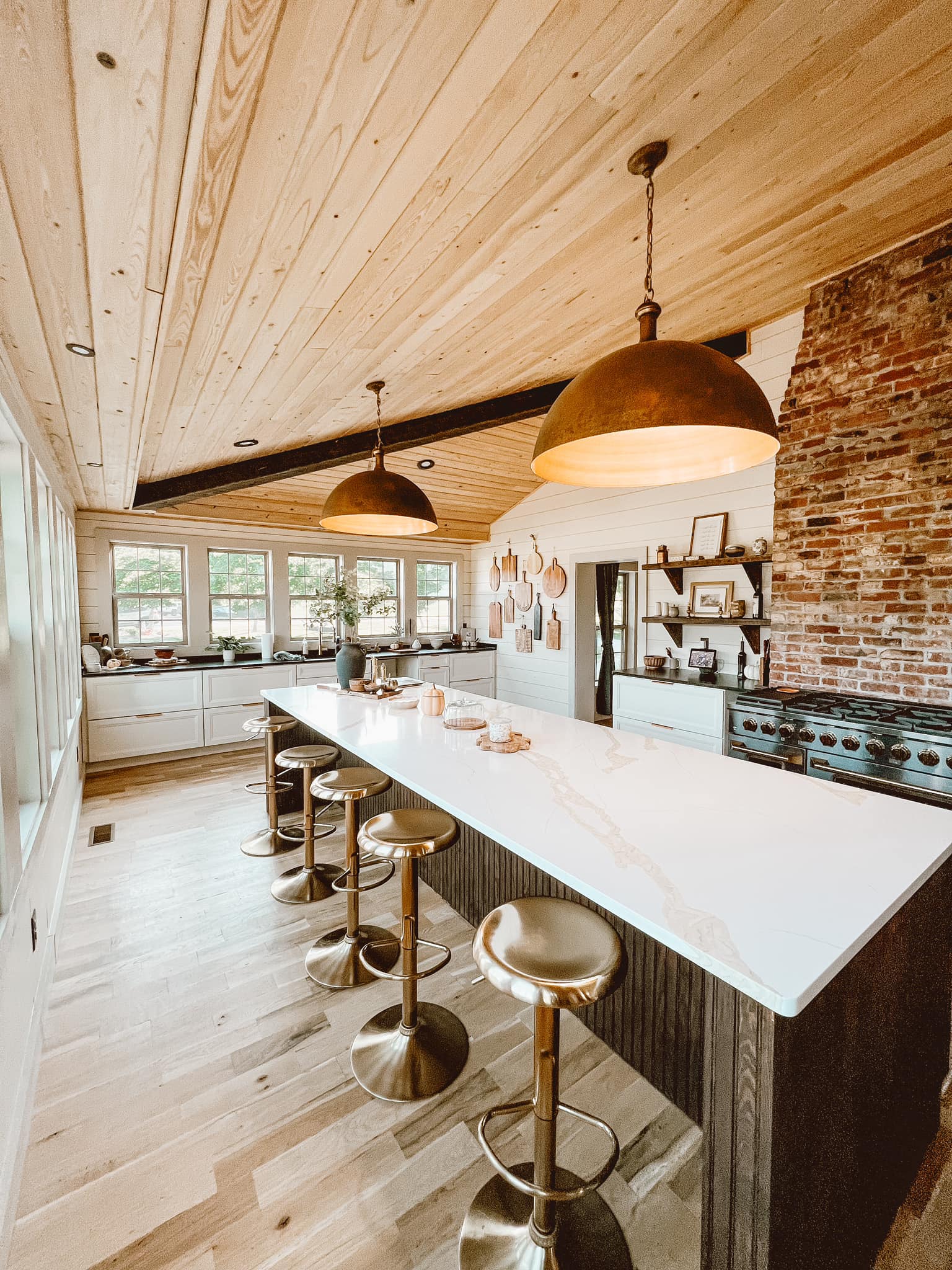 Farmhouse kitchen with a long marble-topped island lined with brass stools, dome pendant lights, white cabinetry, exposed brick, and a wood-paneled vaulted ceiling.