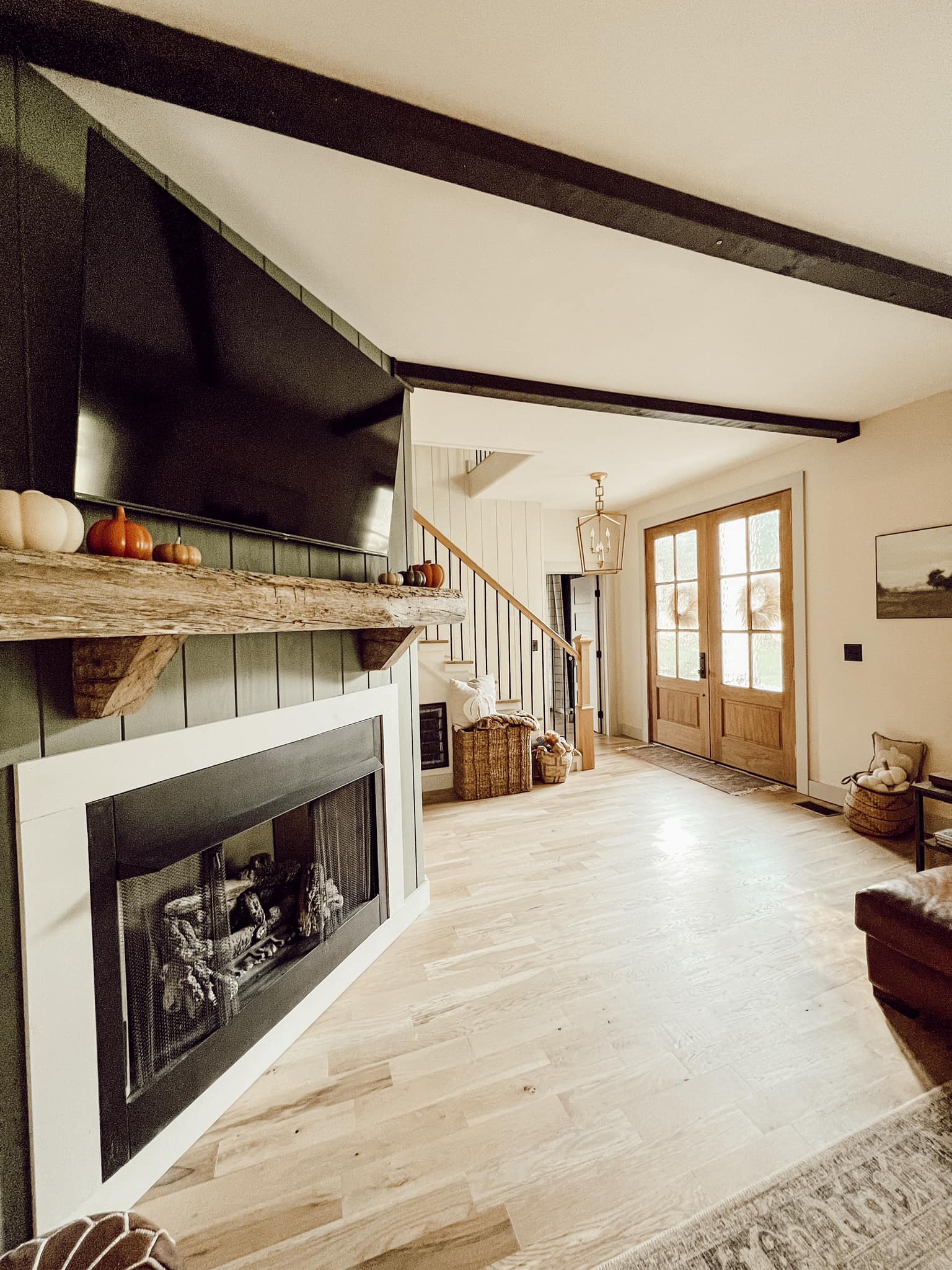 Living room fireplace with a rustic wood mantel decorated with pumpkins, opening to a hallway with a staircase and front entry doors.