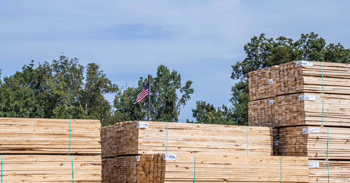 Stacks of bundled lumber outdoors with an American flag and trees in the background.