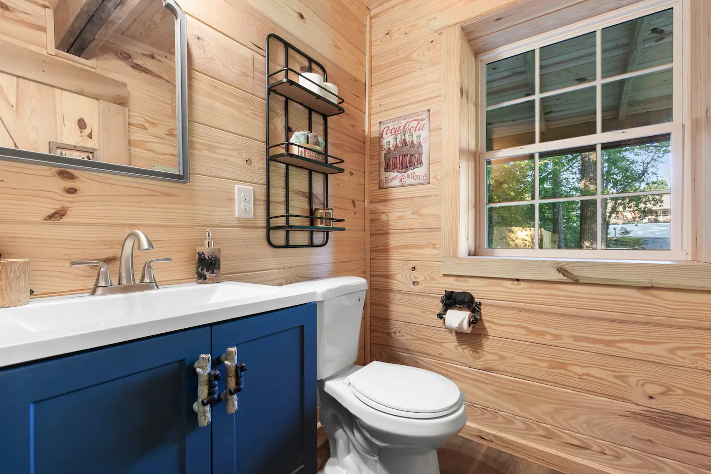 Cabin bathroom with a navy vanity, knotty pine walls, vintage Coca-Cola sign, and a window overlooking trees.