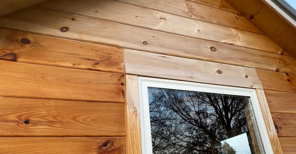 Wood plank siding on a cabin exterior, with some boards showing a darker stain next to unstained boards, framing a window with white trim and tree reflections in the glass.