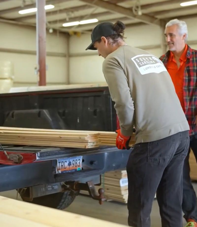 Man in long sleeve gray top and hat loading lumber into the back of a pickup truck
